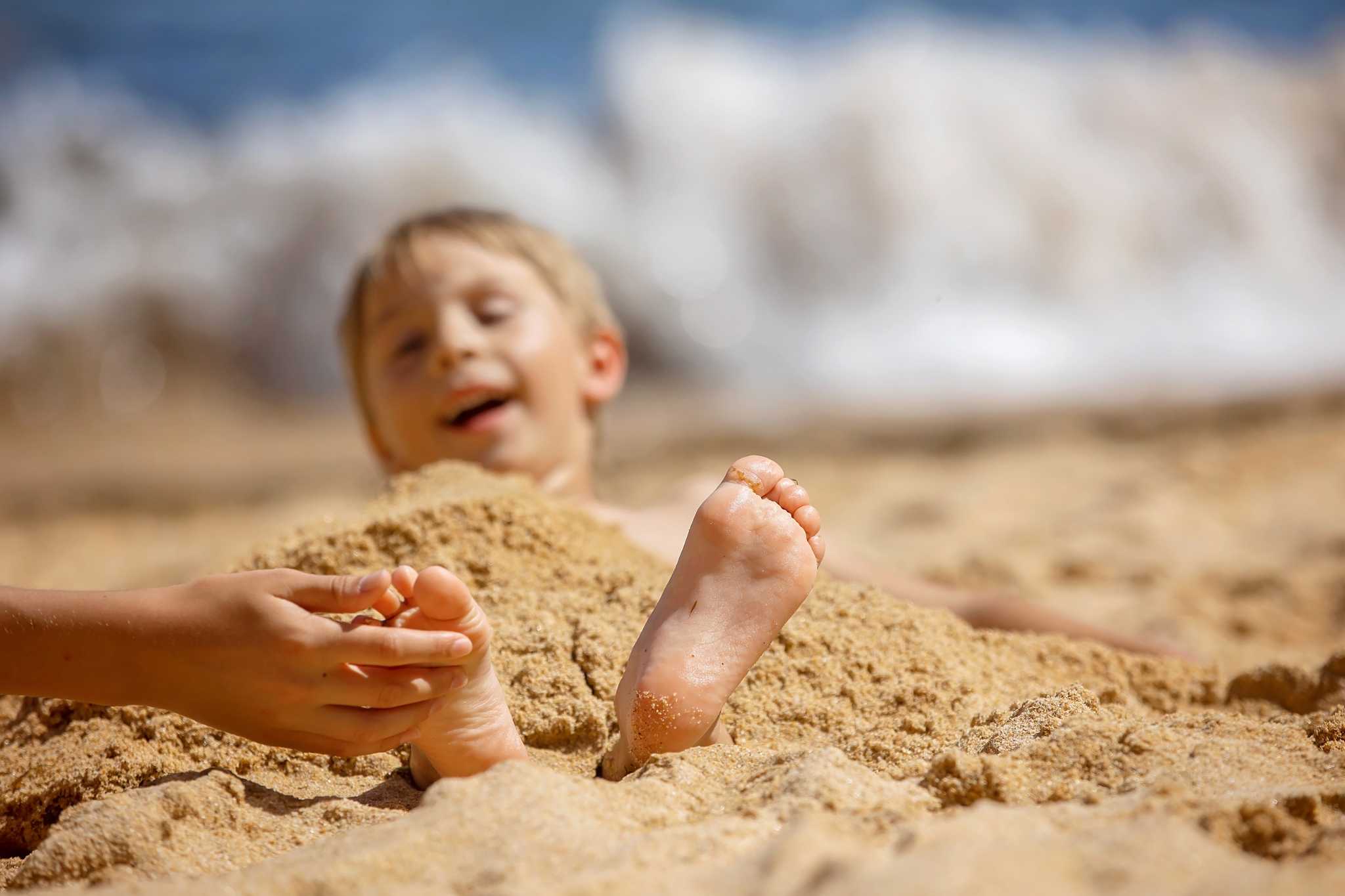 Child, touching sibling on the beach on the feet with feather, kid cover in sand, smiling, laughing
