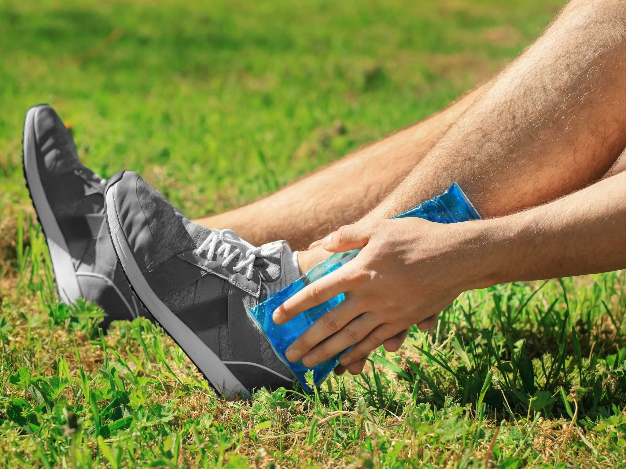 Young man applying cold compress to leg while sitting on grass outdoors, closeup